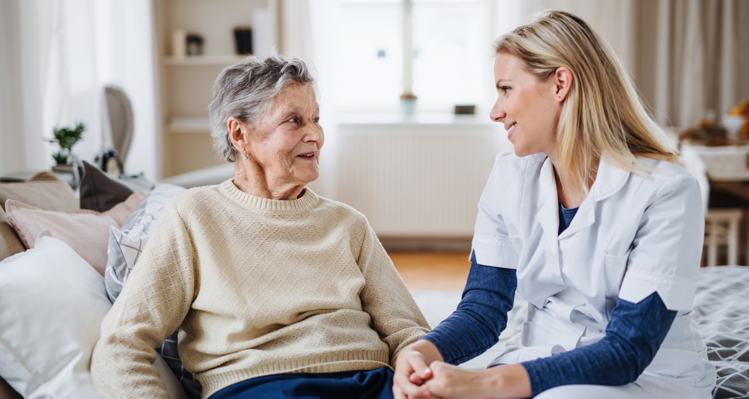 A health visitor talking to a sick senior woman sitting on bed at home.